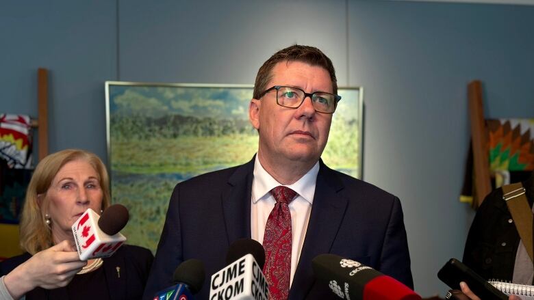 A man in a blue suit, a white shirt and a red tie stands in front of microphones. 