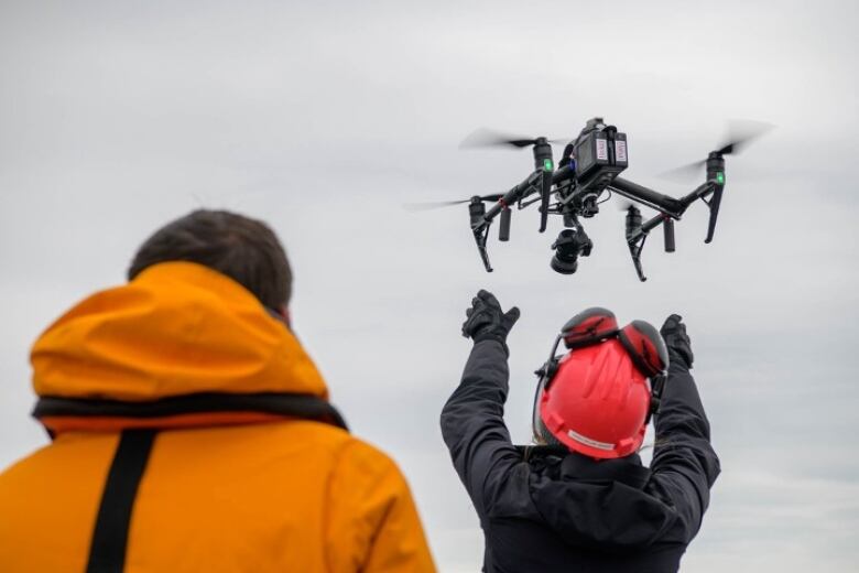 A person in an orange jacket watches as a person in a red hard hat lets a drone fly above their head. 