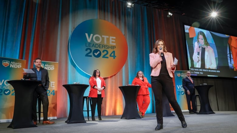 a woman speaks on a stage while others stand by bar-height tables behind her.