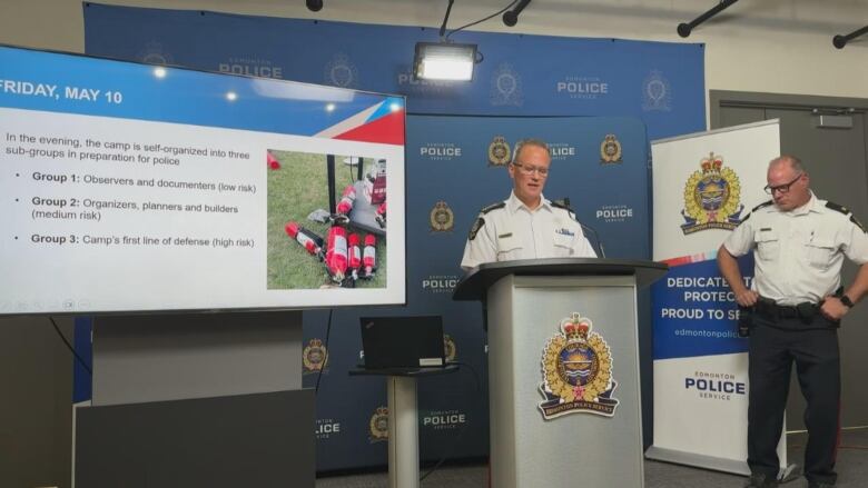 Two police officers stand in a conference room. One of them stands at a podium, speaking into a microphone, while a large screen projects a slideshow presentation to his right.