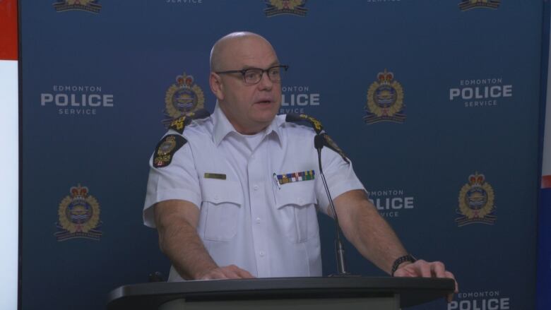 A bald white man, wearing a white short-sleeve button up shirt, is standing in a conference room and speaking at a podium.