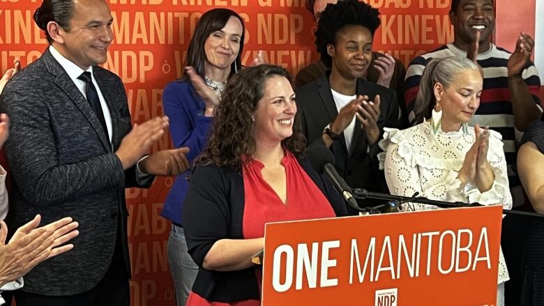 A woman standing at an orange podium smiles as she is surrounded by people clapping. 