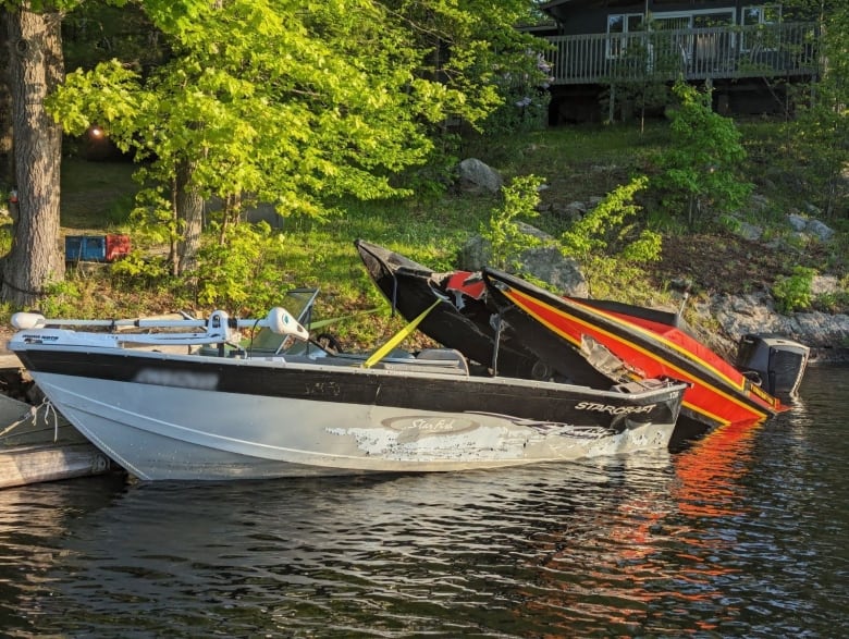 One grey and one orange boat sit in the water next to a grassy shore. Both boats are clearly damaged, and the orange boat is partially on top of the grey one.