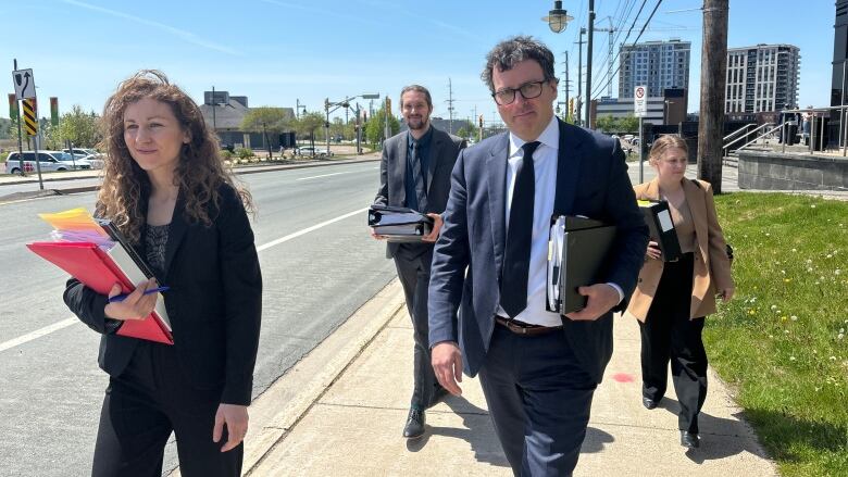 A woman with curly hair holding a binder and a man with files walk ahead of two other people on a sidewalk.