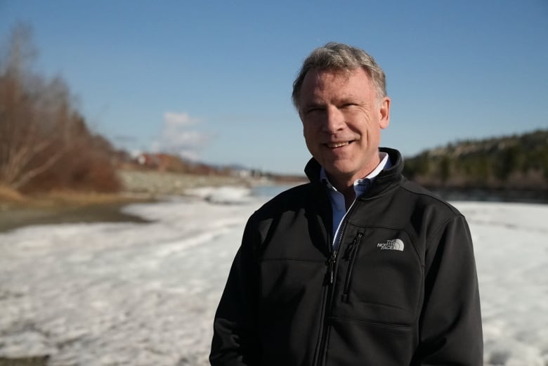 A smiling man stands on the shore of a partially-frozen river.