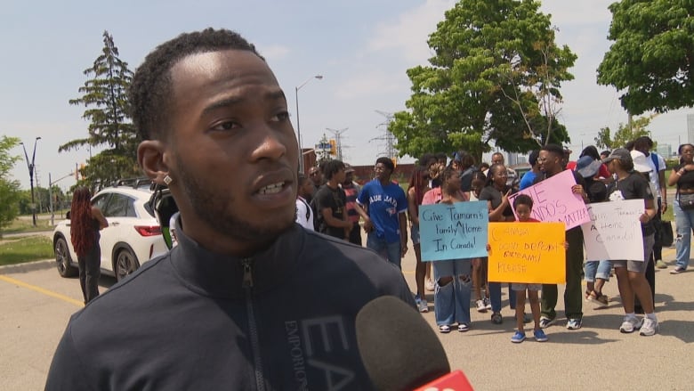 A young man speaks into a reporters microphone in front of a crowd at a rally on a sunny spring day outside a parking lot