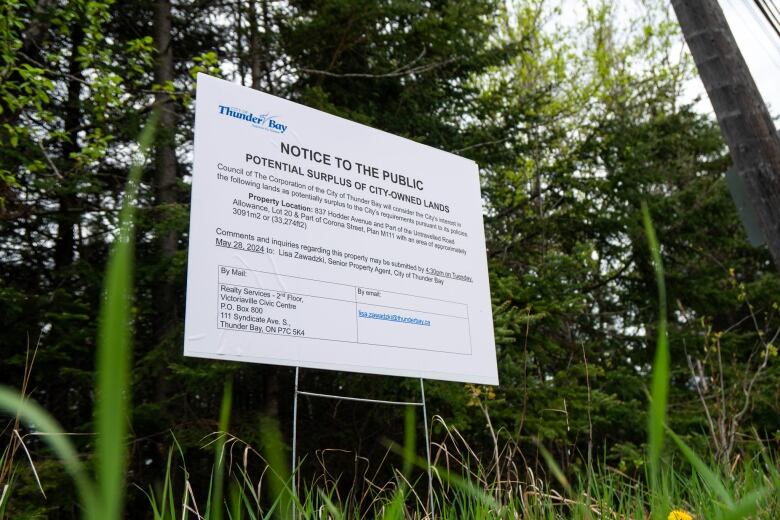 A white sign with black text is seen protruding from green grass against a backdrop of green trees.