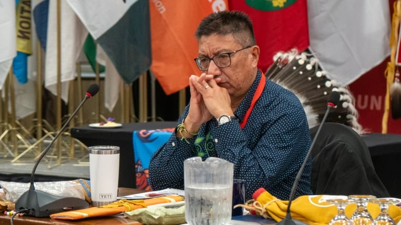 A man wearing glasses and a blue collared shirt sits at a table in front of a microphone. His chin rests on his interlaced hands. 