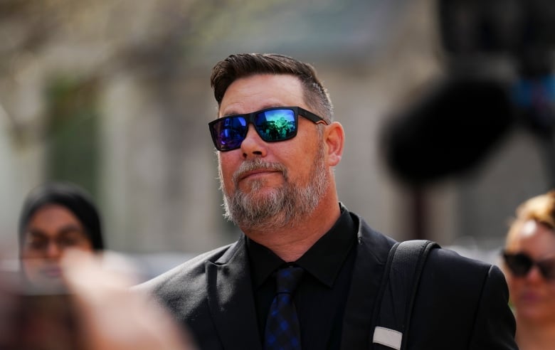 A man in a dark suit and sunglasses outside on a sunny spring day.