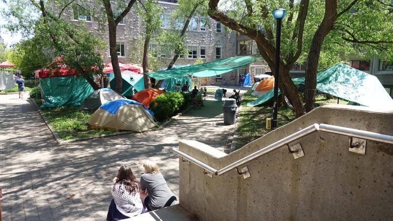 Two women sit on cement steps. In front of them is a large area covered in tents and tarps. It's a sunny day.