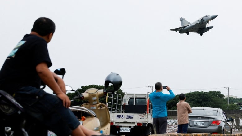 People are shown with their backs to the camera on what appears to be a small bridge looking up at the sky at a fighter jet.