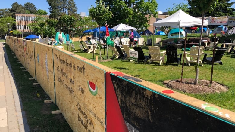 A barricade made of plywood between a sidewalk and a greenspace as part of a protest. There are tents and chairs in the greenspace.