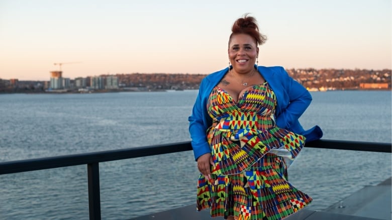 A woman with her hair in a ponytail wears a patterned dress with a blue blazer. She is standing next to the ocean.