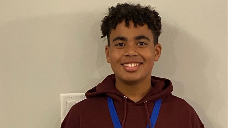 A teen boy wearing a maroon hoodie and blue lanyard stands against a white wall.