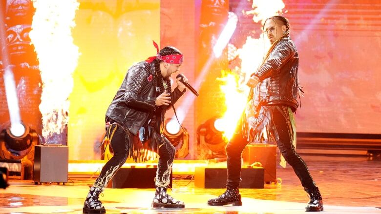 Two men perform on stage with flames in the background.