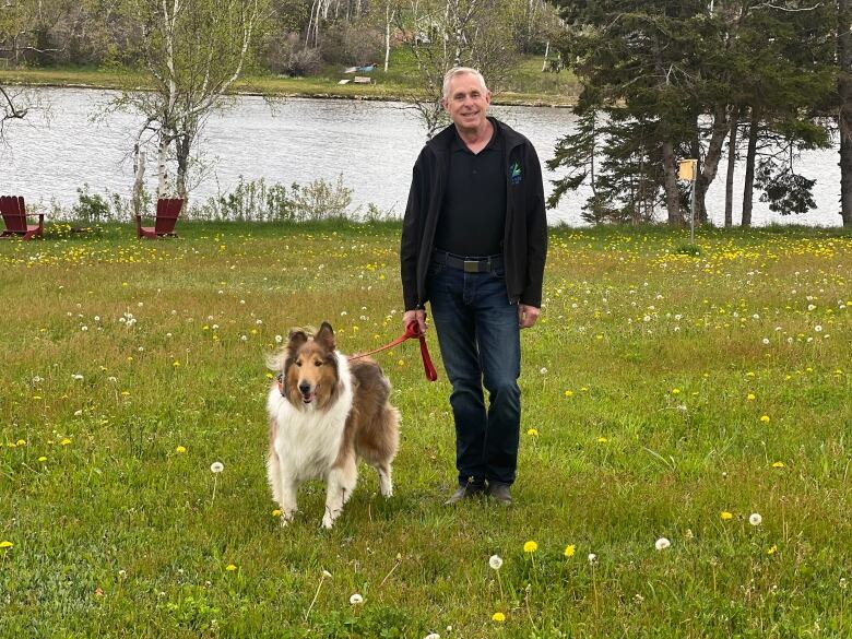 A man with a dog stands in the grass with a river in the background.