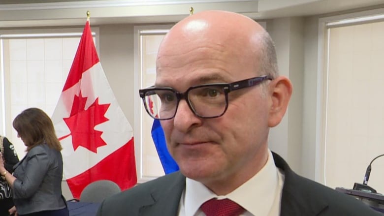 A bald white man is wearing glasses, a red tie, white dress shirt and black suit jacket. He is standing inside a conference room, under fluorescent lights. A Canadian flag stands in the background.