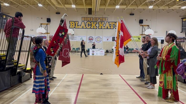 Two lines of people wearing colourful Indigenous clothing hold flags inside a gymnasium. 