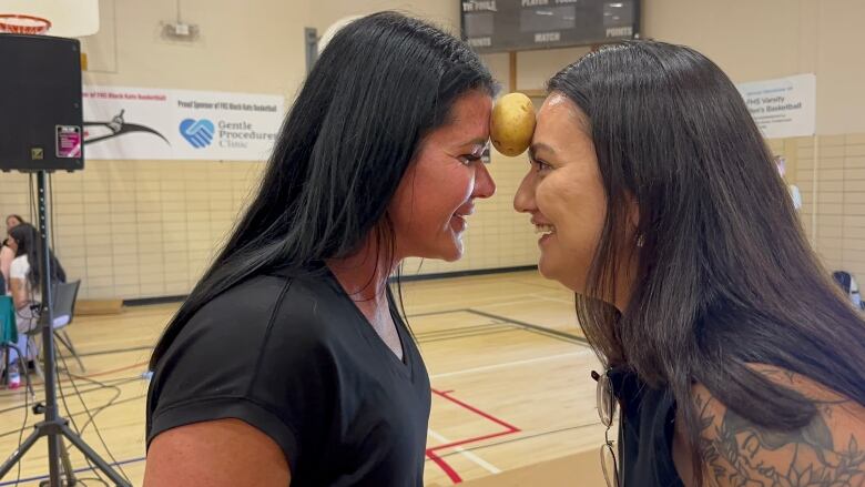 Two women with long, dark hair face each other smiling with a potato between their foreheads.