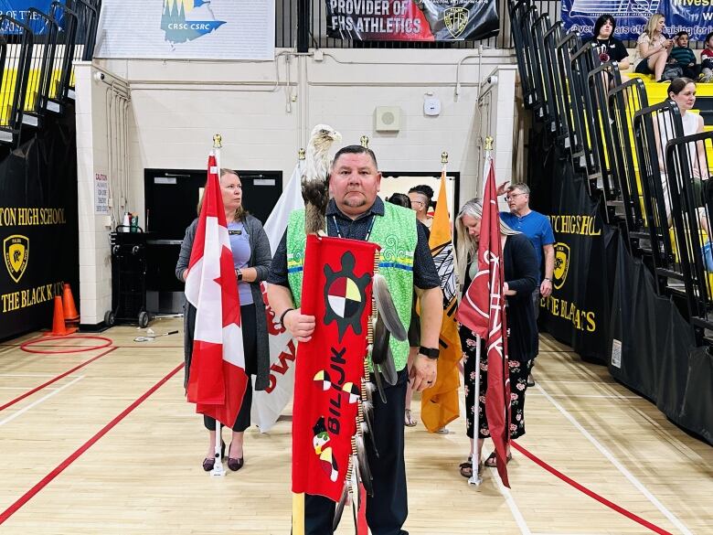 A man wearing colourful, Indigenous clothing stands inside a gymnasium with a faux eagle on his shoulder and holds a flag. More people holding flags line up behind him.