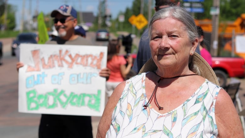 A woman with grey hair with a man holding a sign in the background saying 