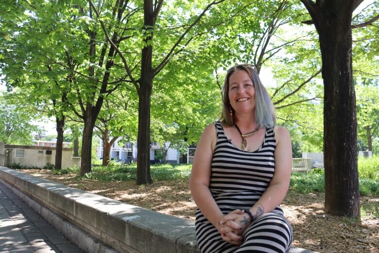 A woman sits on a stone wall, smiling, with trees and sunshine behind her.