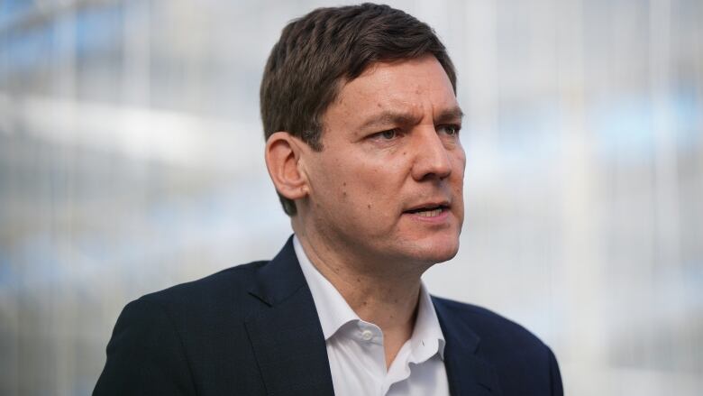 A portrait of B.C. Premier David Eby, a tall man wearing a suit and an open-collared white shirt, speaking during an announcement.