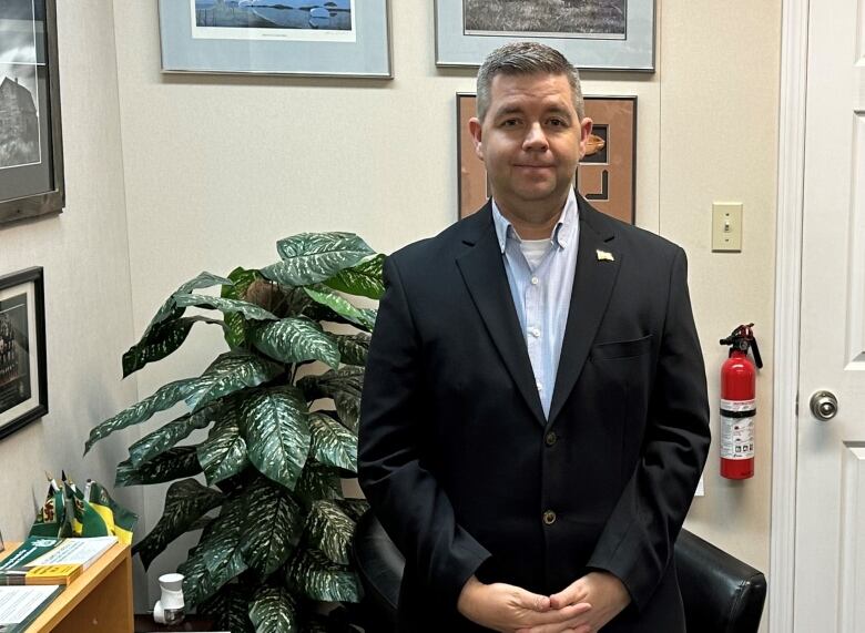 A man wearing a blue button up shirt, grey pants and black suit stands in a room.