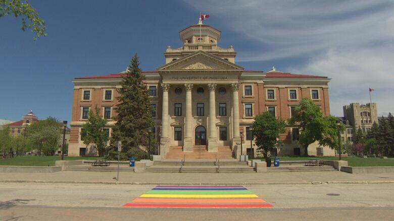 A three-storey brack and tyndall stone building with four pillars in front of its entrance and a rainbow crosswalk in the foreground is pictured.