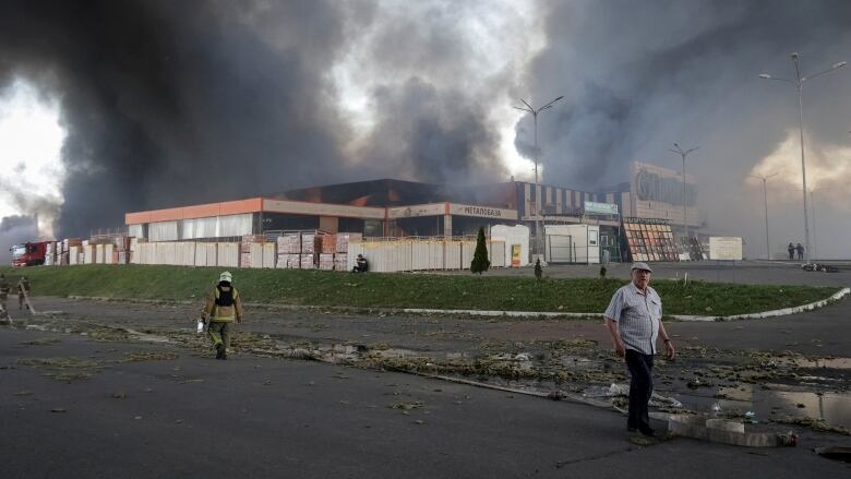 Civilians and firefighters are seen near a commercial structure with smoke billowing from its roof.