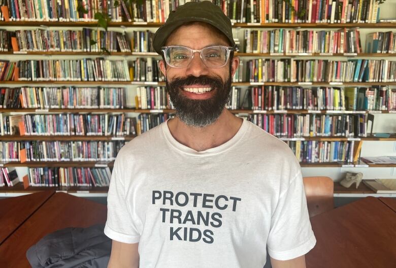 A smiling person stands in front of a very packed bookshelf. They are wearing a hat, glasses and have a thick, dark beard.