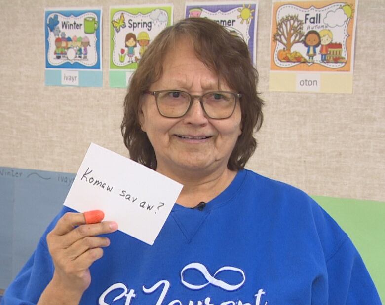 A woman wearing a birght blue sweatshirt with the Mtis infinity symbol holds up a flash card with words in Michif French.