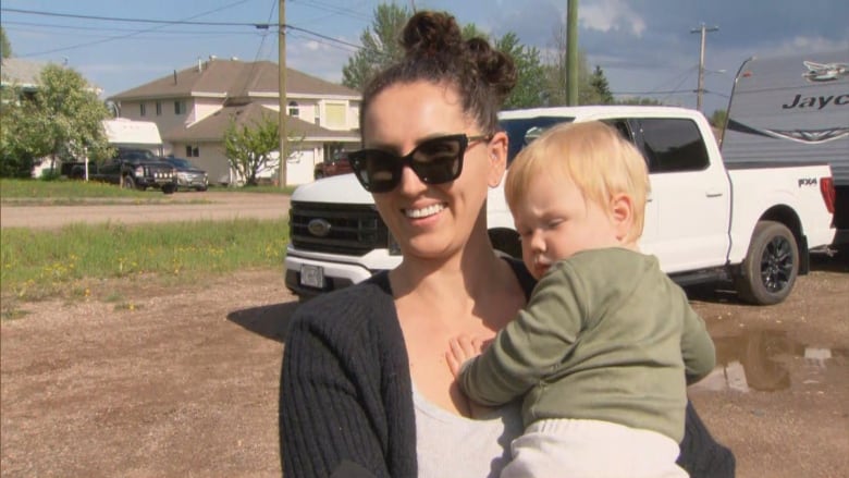 A white woman holding her baby smiles while in a parking lot, with a truck and a house behind her.