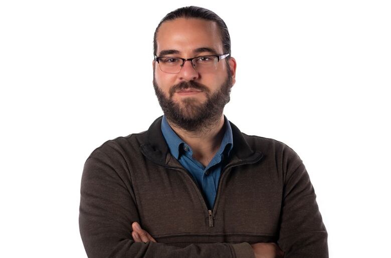 A portrait of a man with brown hair, a beard and glasses, wearing a blue collared shirt and brown sweater, with his arms crossed.