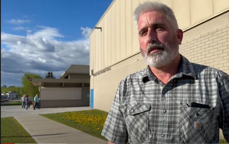 The mayor of Vanderhoof, a man with a white hair and beard and wearing a plaid shirt, has a serious expression on his face as he stands outside a building. 