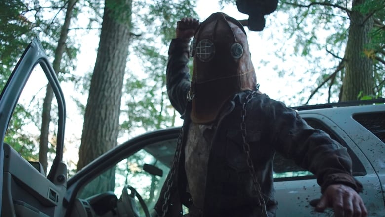 A man wearing a vintage firefighter's mask holds up an axe as if to strike someone. He is standing in front of a car spattered with blood. 