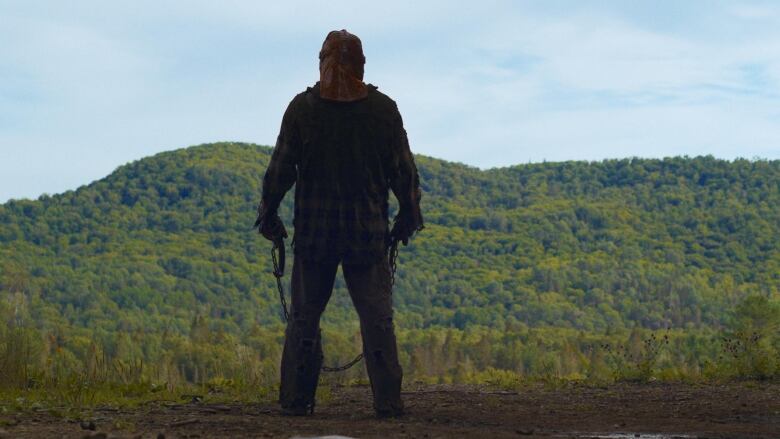 A man wearing a mask and holding a chain looks out at a forested hill.