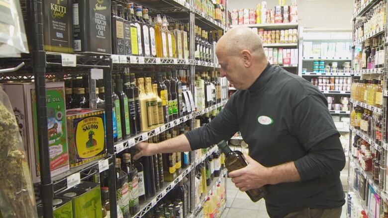 A man stands in a store reaching for a bottle of olive oil. 