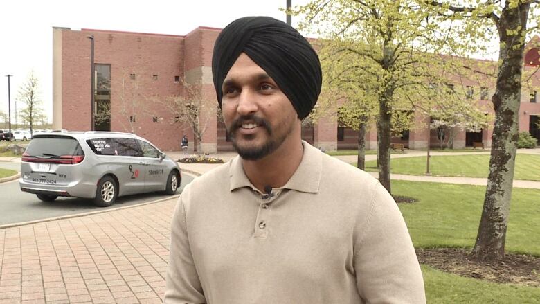 A man wearing a black turban and a khaki shirt standing outside behind a grey shuttle van