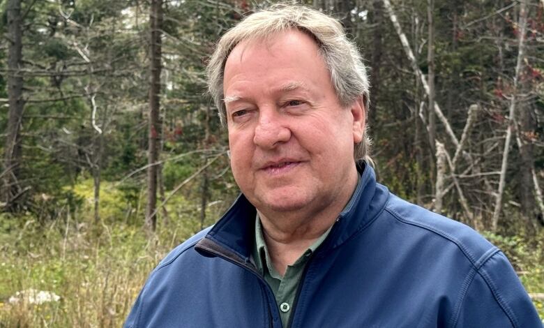 A white man with short greay hair and a navy jacket stands outside, with a wooded area behind him