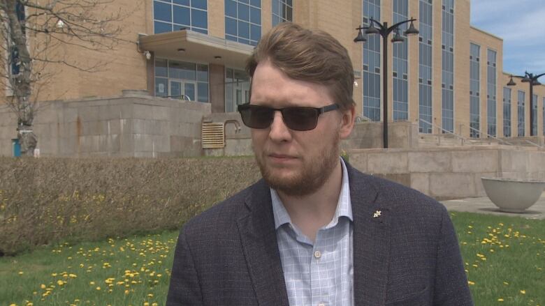 A man wearing sunglasses and a suit stands outside on a grass lawn.