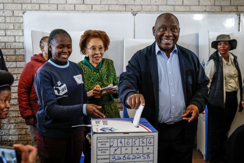 A smiling man puts a ballot into a box, while others look on.