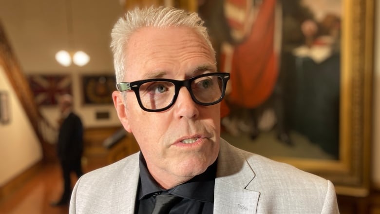 A man with short white hair and dark-framed glasses stands in a hallway and is mid-sentence.