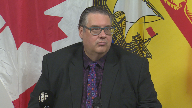 A person in a tie looks at the camera in front of Canada and New Brunswick flags.