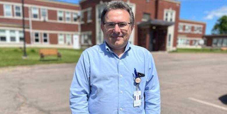 A doctor with a blue shirt stands in the parking lot in front of a brick hospital. 