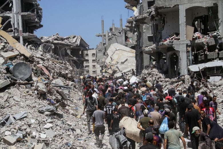 Dozens, possibly hundreds of people, are shown from a distance in an urban setting, with large amounts of concrete debris and damaged buildings shown on either side of them.