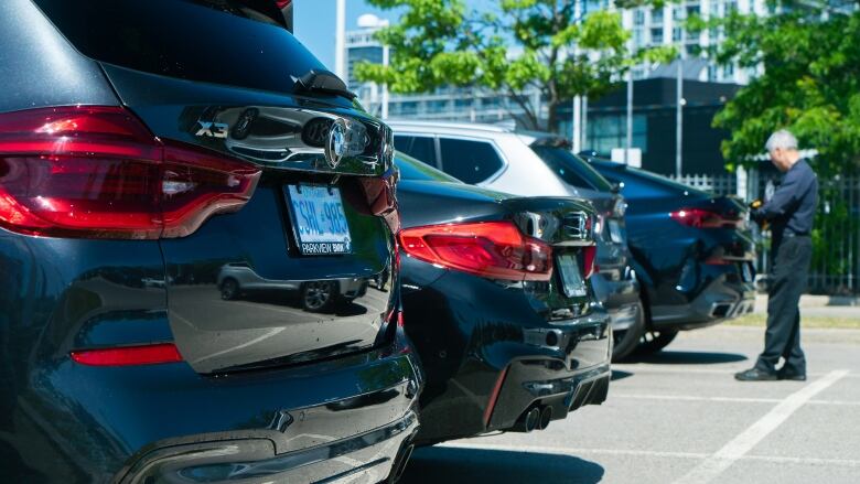 A man stands near the end of a line of several cars in a row in bright sunshine with a tree in the background. 