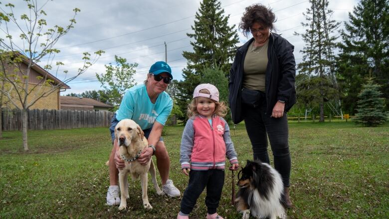 Two adults, a young child and two dogs stand on a patch of green grass.