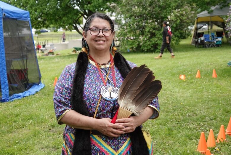 An Indigenous woman in her regalia stands on a grass field, flanked by tents and pylons.