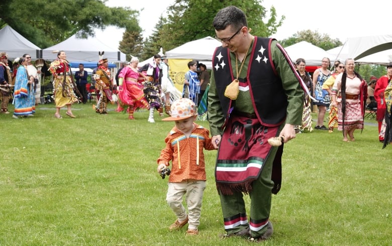 A man holds the hand of a young boy as they dance during a powow. Many other people are following in a line behind them, dressed in Indigenous regalia.